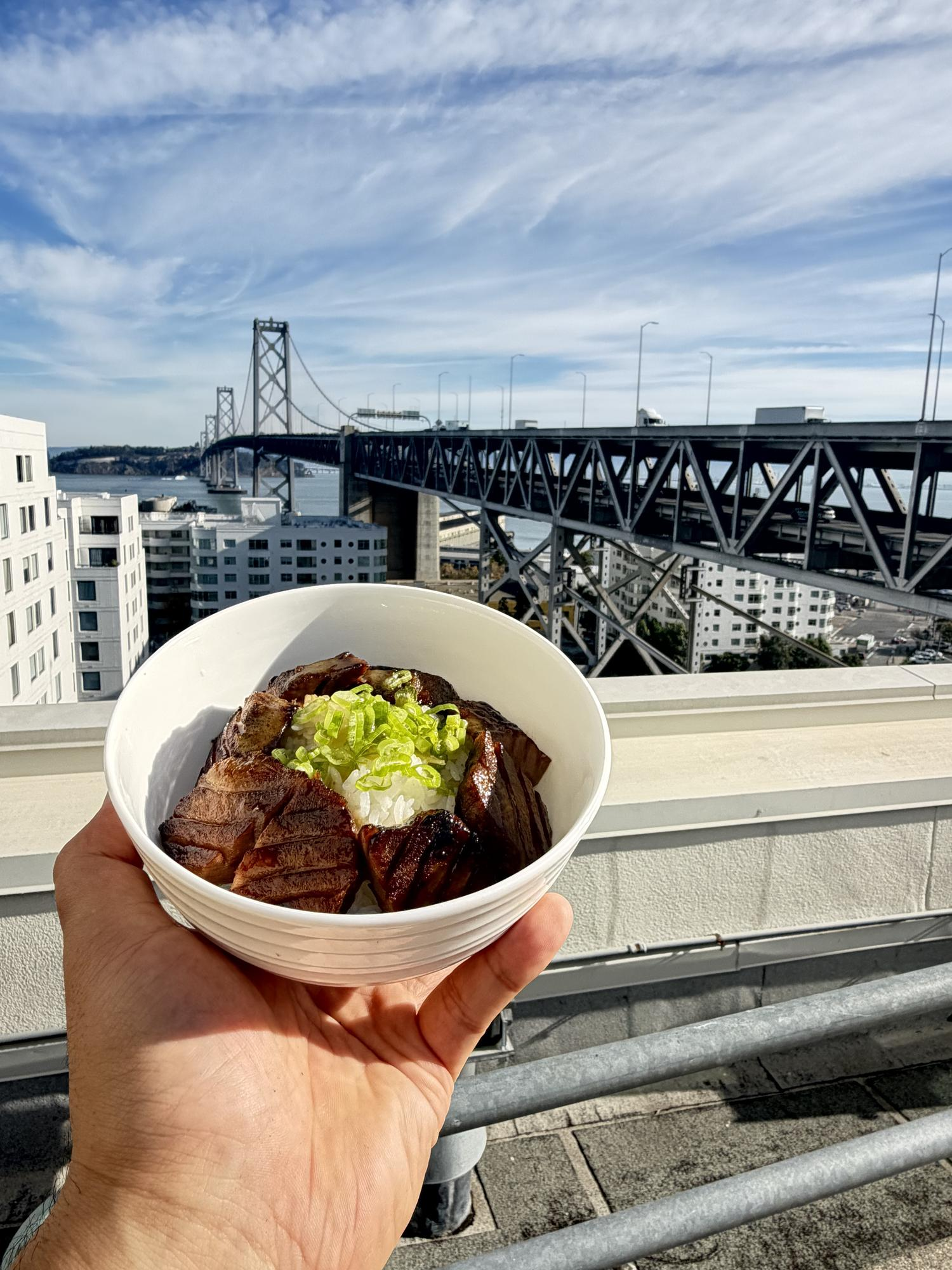A hand holds a bowl of beef and rice with green onions, with the Bay Bridge and city skyline in the background.