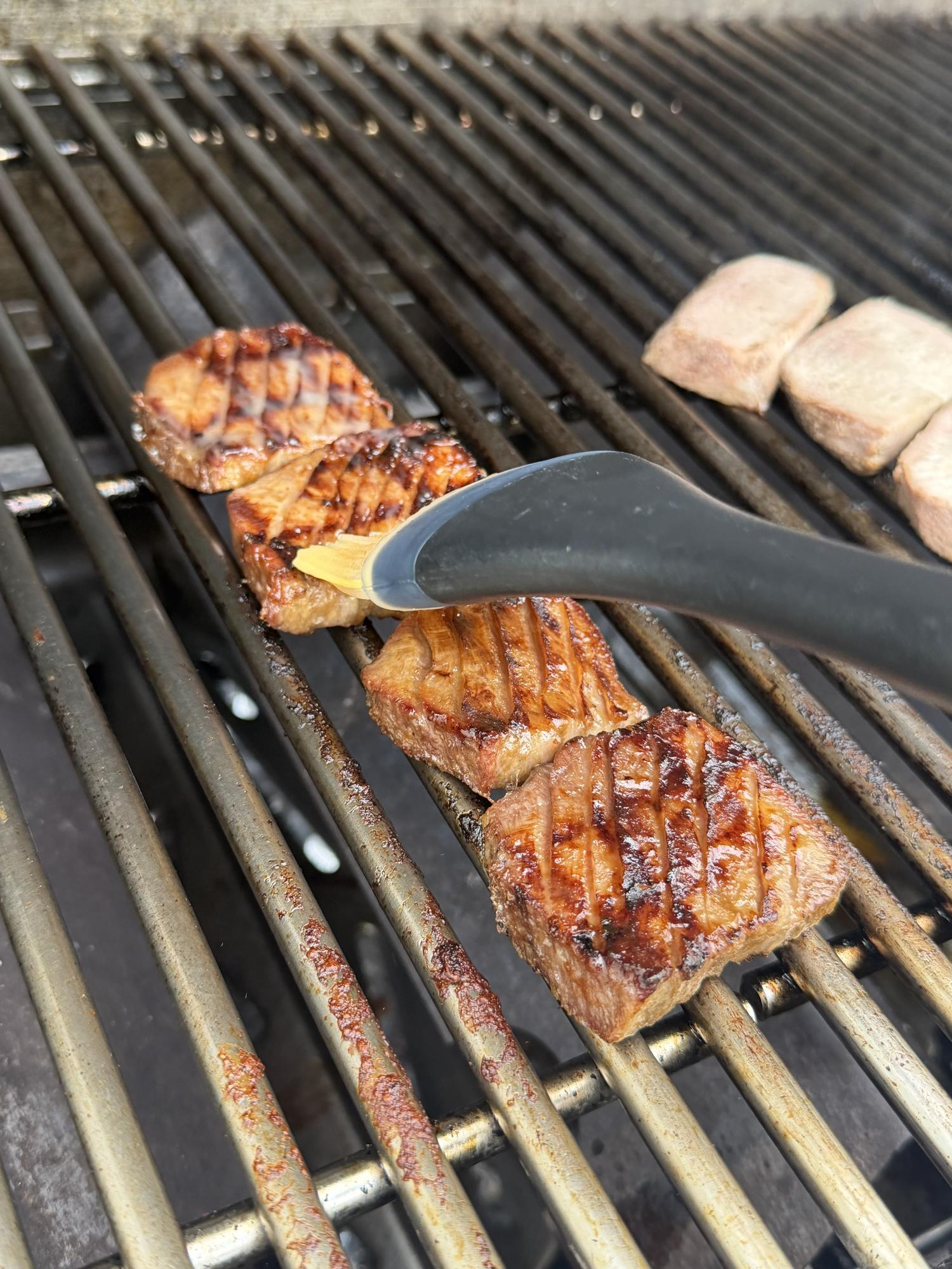 Grilled meat with distinct char marks being basted with sauce on a barbecue grill using a silicone brush.
