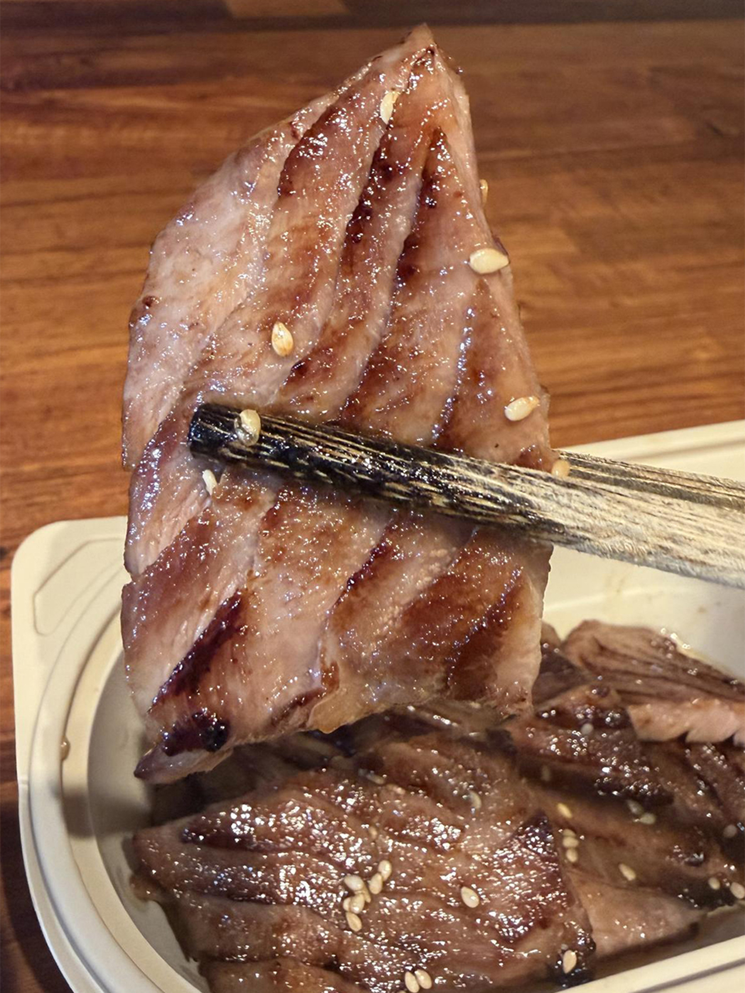 Grilled meat slices with sesame seeds, held by chopsticks over a takeout container.