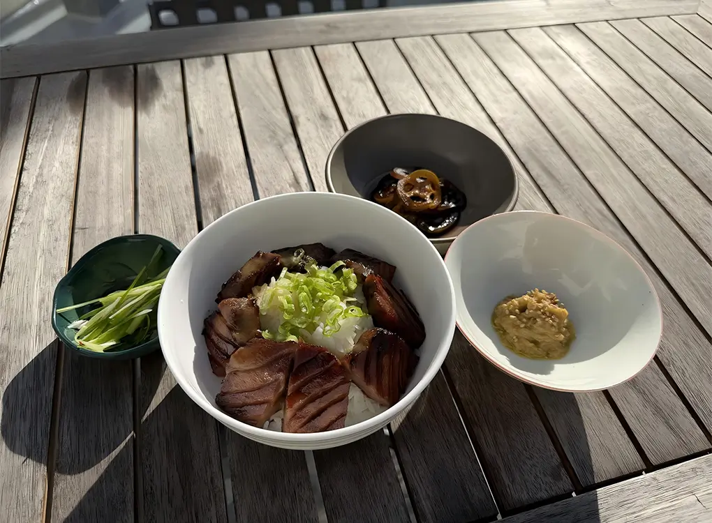 Japanese butadon rice bowl with grilled glazed pork slices topped with chopped green onions, served with side dishes including pickled vegetables, miso paste, and scallions.