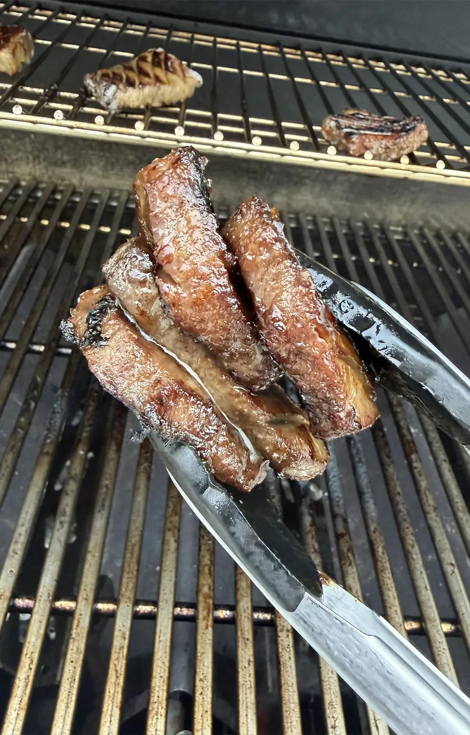 Glazed pork slices being brushed with sauce on a barbecue grill, with raw pieces nearby.