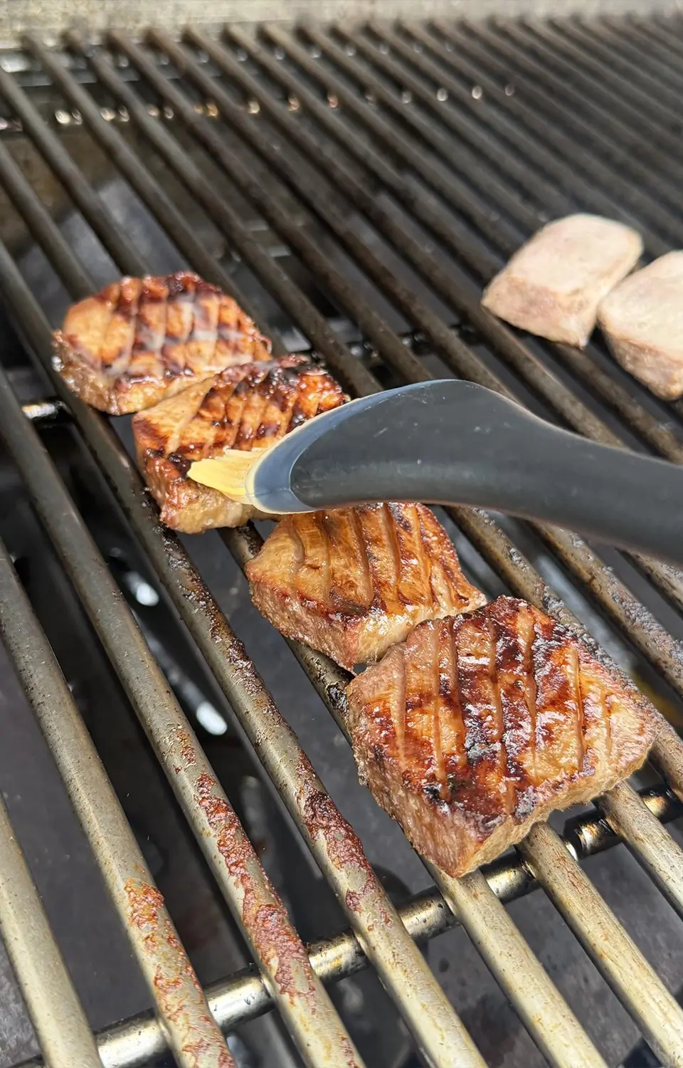 Close-up of tongs holding juicy grilled glazed pork slices over a barbecue grill.
