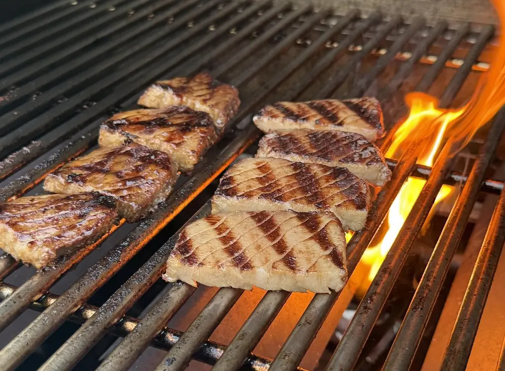 Sliced glazed pork pieces grilling on a barbecue with visible flames and grill marks.