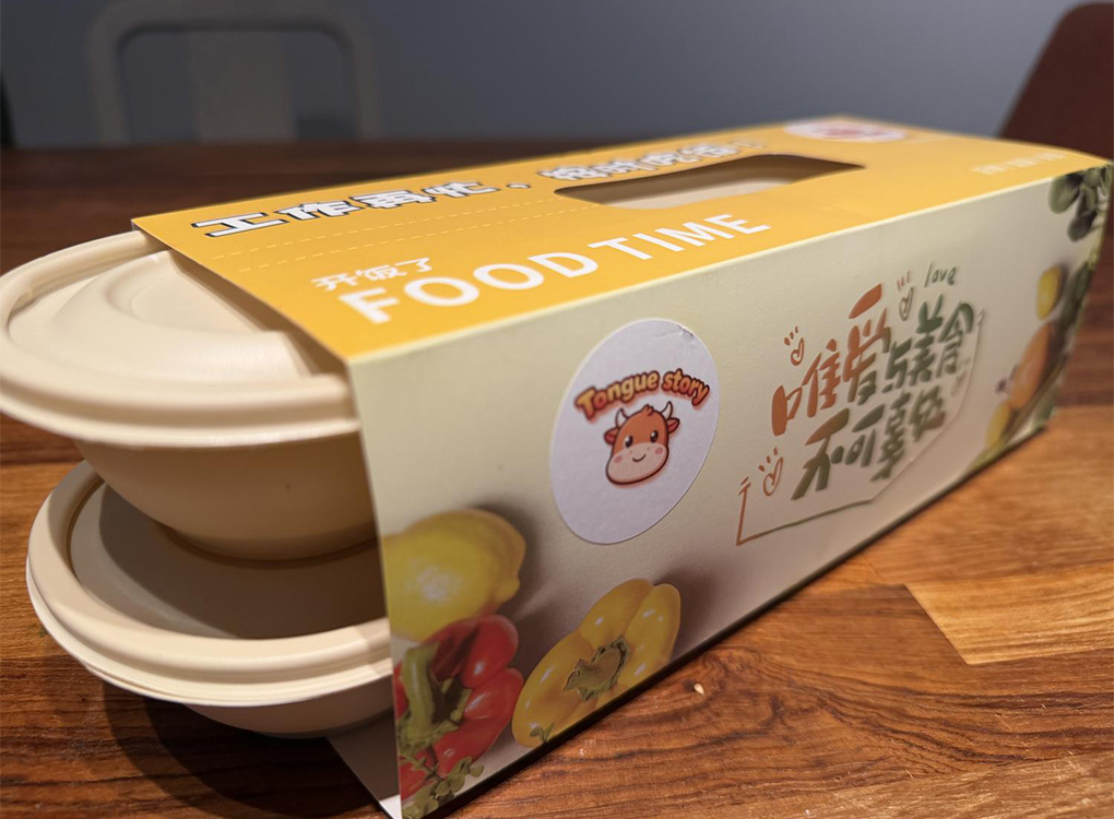A "Tongue Story" takeout box with two bowls, featuring Chinese text and colorful peppers on a wooden table.