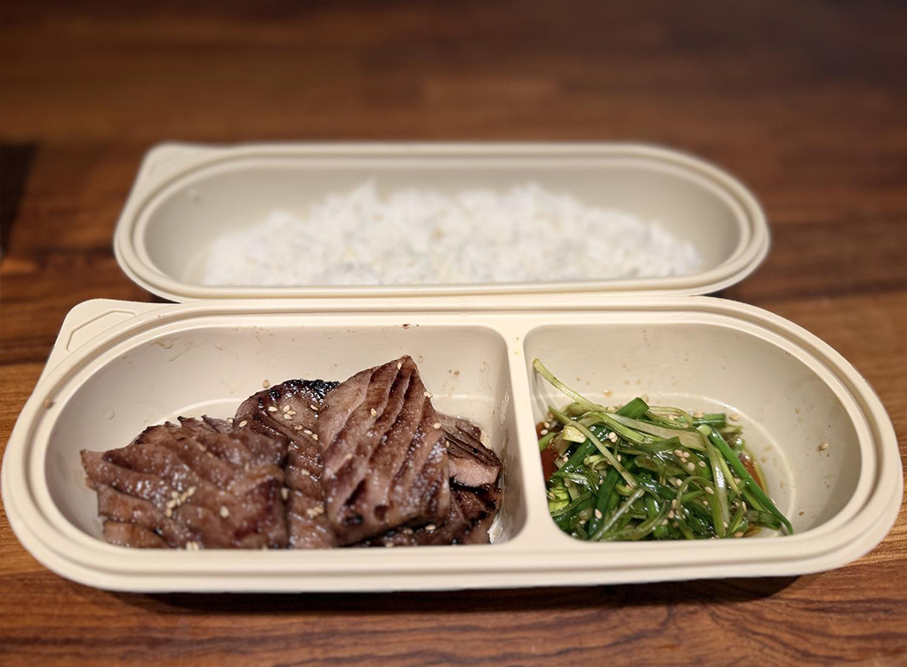 A takeout bento box with grilled meat, green onions, and white rice, placed on a wooden table.